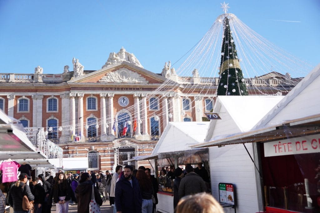 Vue sur le marché de Noël de la place du Capitole à Toulouse, entre chalets blancs, grand sapin décoré et façade illuminée du Capitole.